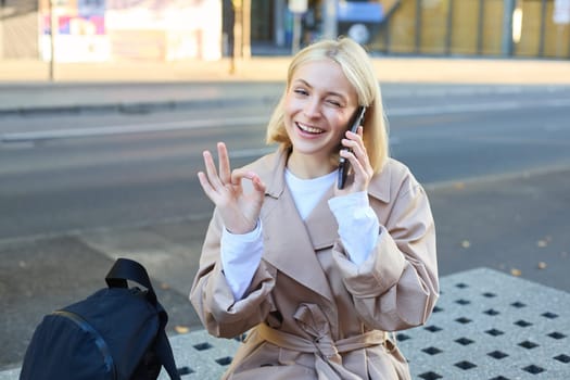Image of young woman talking on mobile phone, sitting on bench outside, showing okay, ok approval gesture, made a deal over the telephone.
