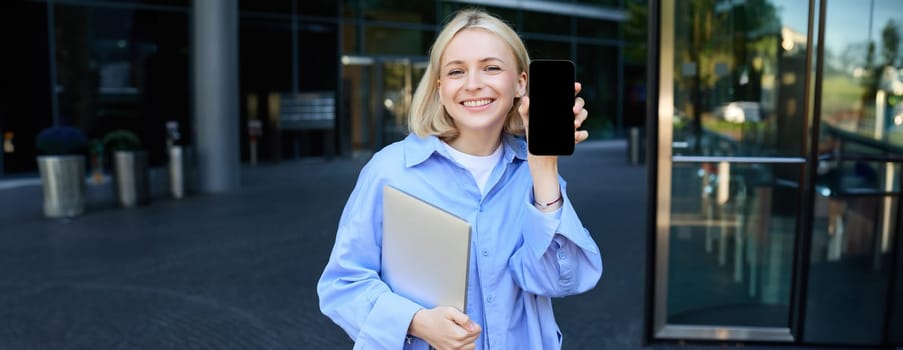 Portrait of stylish, modern young office worker, woman showing her smartphone screen, mobile phone app on display, posing near campus or office building, holding laptop.