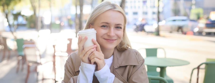 Close up of cheerful blond woman, holding cup of coffee, drinking tea in street outdoor cafe, smiling with happy face expression.