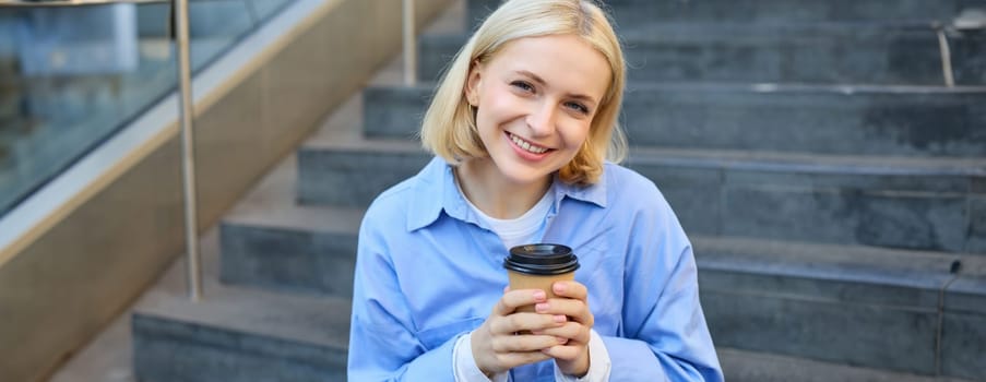 Close up portrait of female model, cute student with cup of coffee, sitting on stairs near campus, spending time outside.