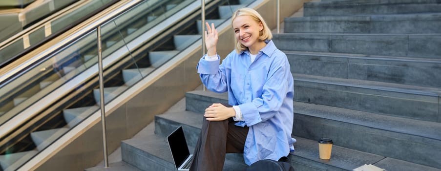 Friendly student girl, saying hi, waving hand at someone, sitting on stairs and drinking coffee, using laptop computer and public wifi.