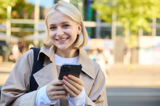 Close up portrait of smiling, beautiful blond girl, holding backpack and mobile phone, standing on street with lots of sunlight, buildings and road behind, looking happy at camera.
