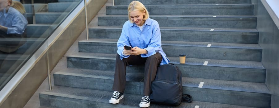 Portrait of young modern woman, student with backpack, sitting on city stairs on lunch break, using smartphone, looking aside.