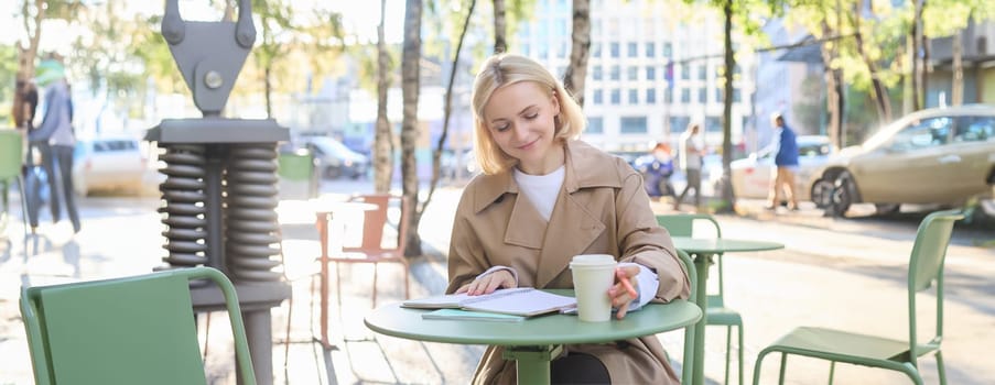 Portrait of young smiling woman, working outdoors, sitting with notebook in cafe and writing, drinking coffee, student doing homework on fresh air in relaxing atmosphere.