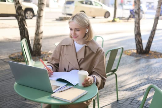 Lifestyle and people concept. Woman in outdoor cafe, sitting with laptop on street, connects to online course or lecture, doing homework, making notes, wearing wireless headphones, drinking coffee.