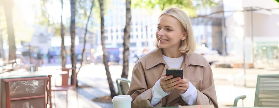 Portrait of blond smiling woman sitting in cafe, enjoying cup of takeaway coffee, using mobile phone, working on smartphone, social media blogger making post online.