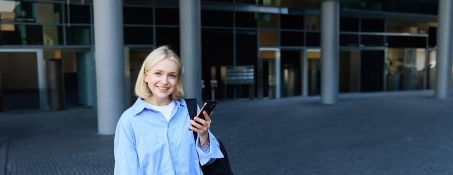 University and people concept. Vertical portrait of young woman standing near office building on street, student in college campus, holding smartphone, laptop and backpack on shoulder.