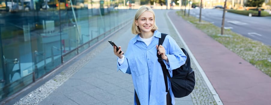 Lifestyle portrait of smiling blond woman with smartphone, walking on street with backpack and looking happy, on her way to college, using mobile phone.