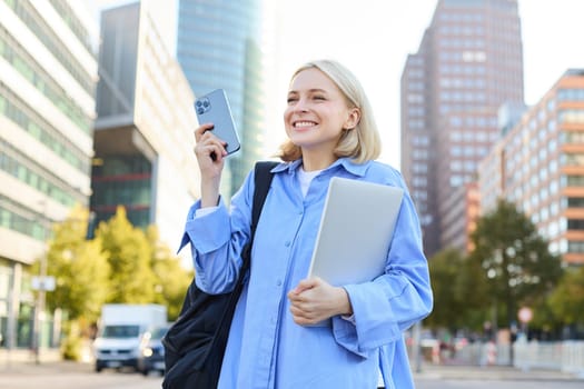 Portrait of happy, excited young woman, standing in big city with smartphone and laptop, looking amazed and inspired, walking along the street.