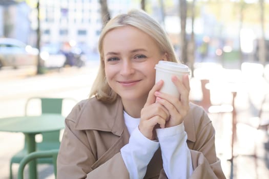 Close up of cheerful blond woman, holding cup of coffee, drinking tea in street outdoor cafe, smiling with happy face expression.