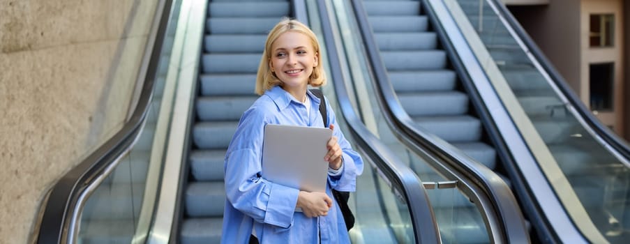 Lifestyle portrait of young woman with laptop, walking away from escalator, heading to tube metro, smiling and looking aside.