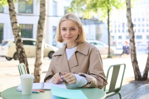 Portrait of young stylish student, woman working outdoors in cafe, doing homework, writing essay, holding markers, smiling at camera.