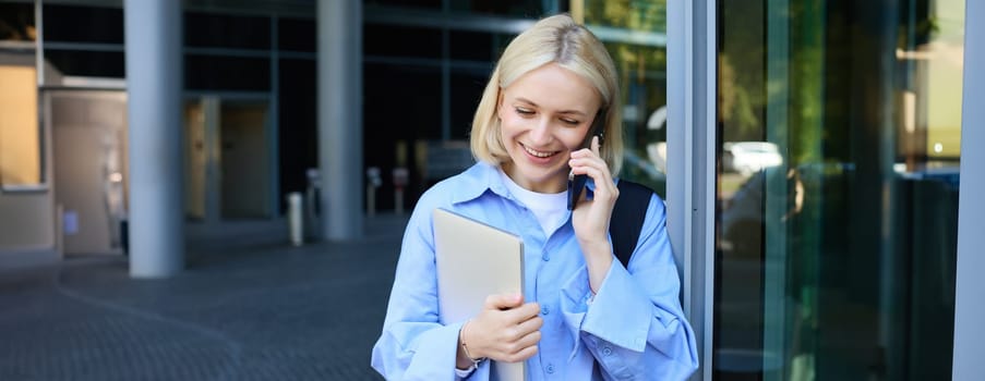 Close up portrait of smiling blond woman, holding laptop, has backpack on shoulder, talking on smartphone, answer a phone call.
