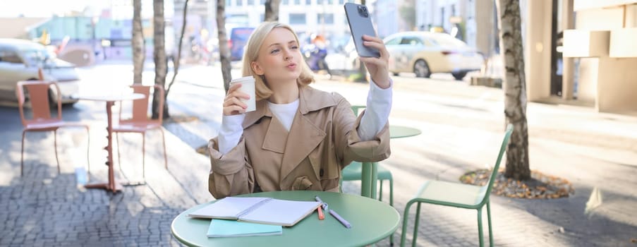 Fashionable young blond woman, taking selfie with favourite drink in coffee shop, puckers lips, creating content for social media, influencer making photos.