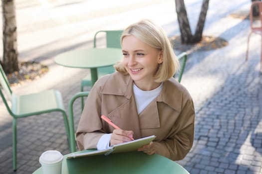 Image of young smiling woman, sitting in outdoor cafe, doing sketches, drawing art on street, spending time on coffee shop and creating art.