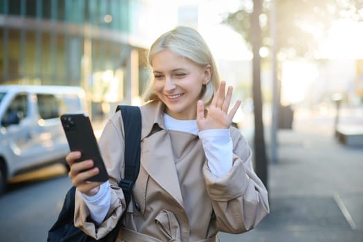 Image of happy, beautiful young woman on street, answers video call, waves hand at smartphone camera, says hello, talks to friend over the mobile app, walking along road.