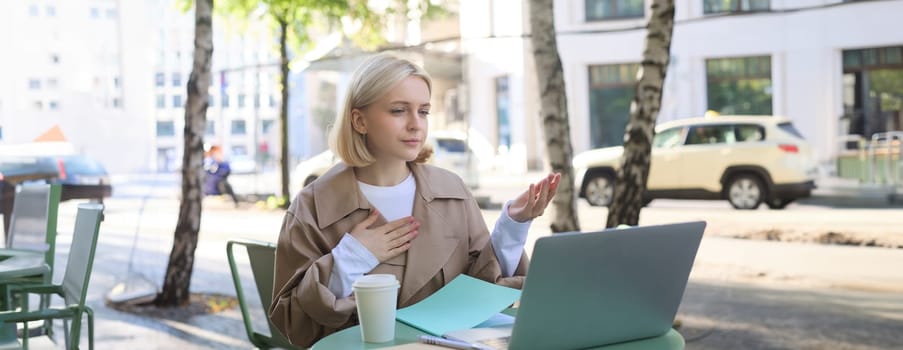 Image of young stylish woman, sitting on street, drinking coffee in cafe, working remotely on laptop, attends online course or doing homework.