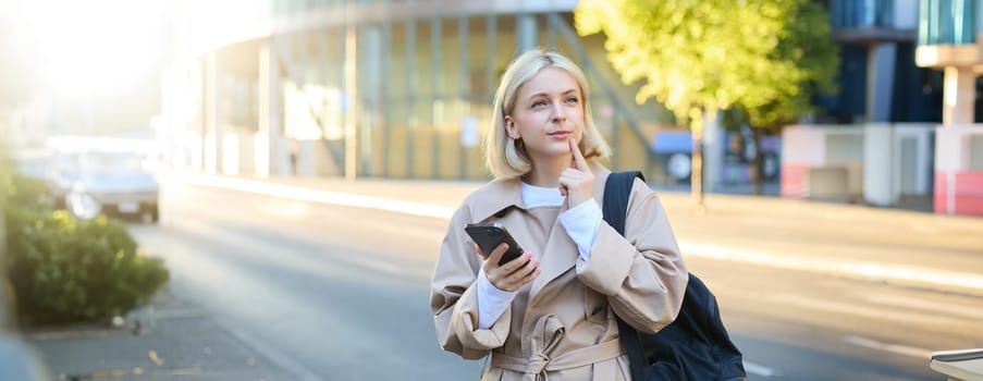 Portrait of woman with thinking face, standing with backpack on street, holding smartphone, pondering, making a decision with thoughtful face.