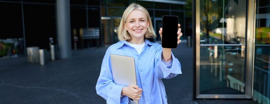 Portrait of beautiful young woman, student posing on street near campus, holding laptop, showing mobile phone screen, demonstrating app or promo on smartphone and smiling.