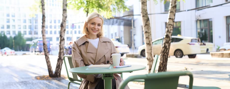 Portrait of young female student, woman with notebook, sitting in cafe and drinking coffee, smiling happily.