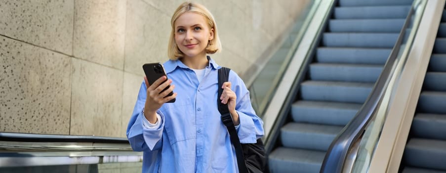 Portrait of young college student, woman with backpack and mobile phone, standing near escalator, reading notification on smartphone.