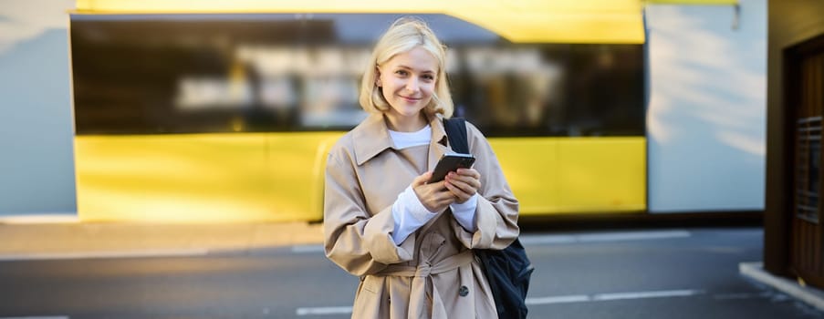 Portrait of modern young woman, student using smartphone on street, bus behind her, smiling and looking happy at camera.