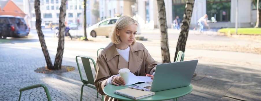 Image of young modern woman, sitting in cafe outdoors, drinking coffee or tea, looking at laptop, working on project, freelancing, making notes in notebook. Lifestyle concept