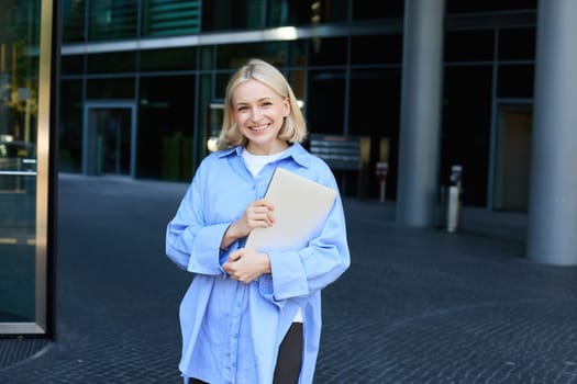 Portrait of young blond woman, student standing near her campus with notebooks and documents, wearing blue shirt and smiling at camera. Education concept