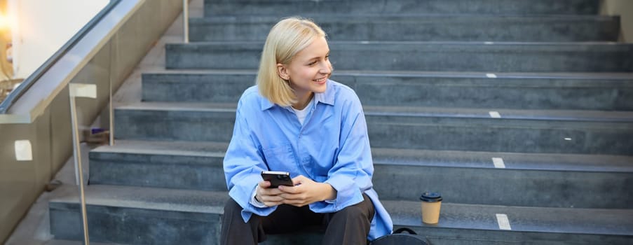 Portrait of young modern woman, student with backpack, sitting on city stairs on lunch break, using smartphone, looking aside.
