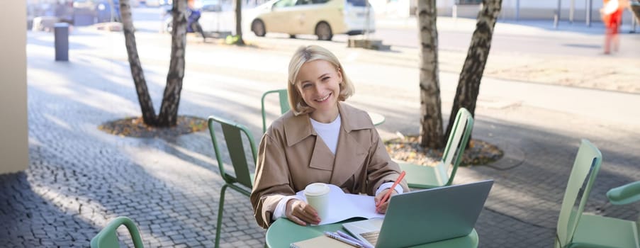 Image of young blond modern woman, sitting with laptop outside in cafe, drinking coffee drink and working on project, using computer.