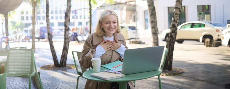 Image of young smiling woman working remotely, connects to online call, chatting via laptop, looking pleased, feeling grateful, laughing, sitting in outdoor cafe.
