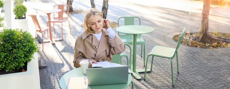 Lifestyle and people concept. Woman in outdoor cafe, sitting with laptop on street, connects to online course or lecture, doing homework, making notes, wearing wireless headphones, drinking coffee.