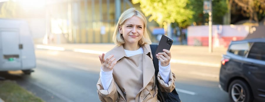 Portrait of confused blonde woman on street, carries backpack, holding smartphone, frowning and looking puzzled, unsure where to go, using mobile map.