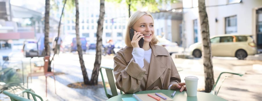 Close up of cheerful blond girl, woman talking on mobile phone, sitting in city centre, outdoor cafe, taking break from work or studying, chatting on telephone.