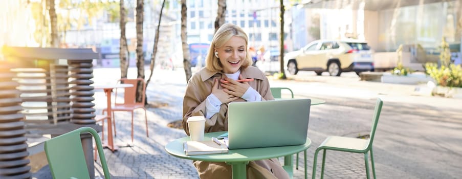 Portrait of young blond woman with laptop, sitting on street, drinking coffee in outdoor cafe, working, wearing wireless headphones, doing homework, freelancing.