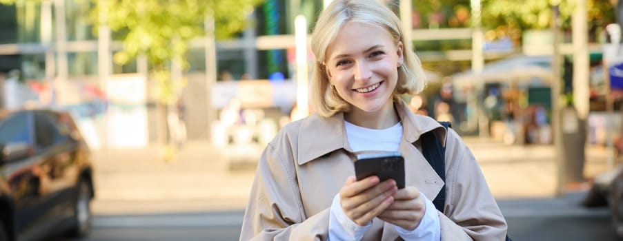 Close up portrait of young smiling blond woman, student on street, using mobile phone, checking messages on smartphone, walking on road.