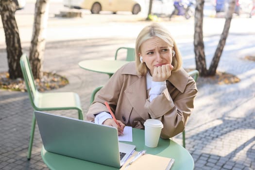 Lifestyle and technology concept. Young blond working woman in trench coat, studying, working on project on laptop, sitting in coffee shop on sunny day, making notes, attending online course.