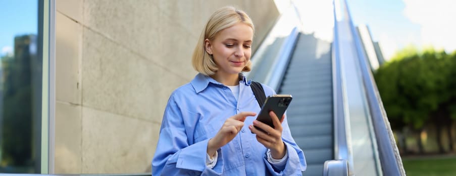 Close up portrait of young blond woman in city centre, standing near escalator, checking her mobile phone, reading message on smartphone. Lifestyle concept