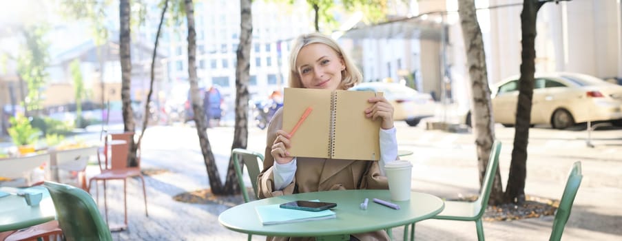 Outdoor shot of young beautiful blond girl with notebook, woman sitting in outdoor cafe, writing in journal, making plans in her planner, putting notes.