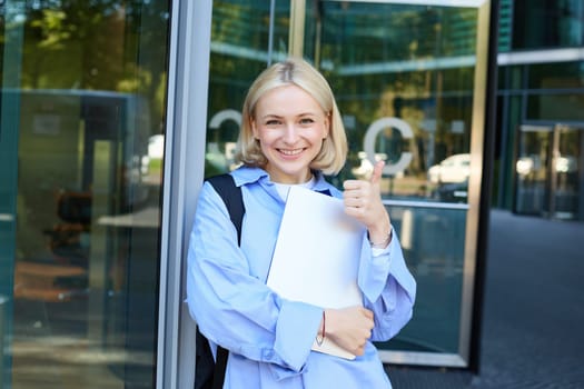 Image of young college girl, young woman with laptop, posing near university building, standing on street, smiling, concept of education and people.