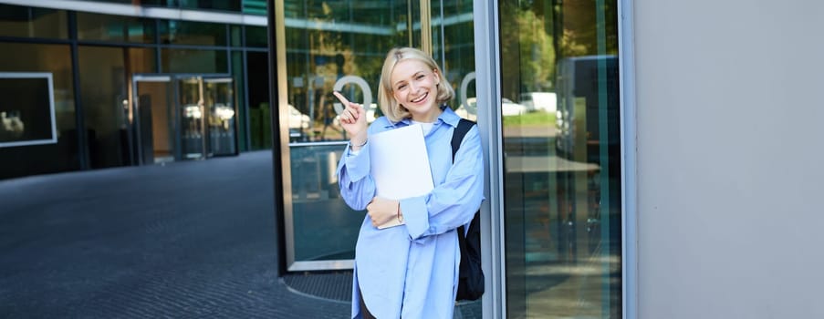 Portrait of smiling young woman, student or office worker with laptop, pointing finger at upper right corner, showing building and looking happy.