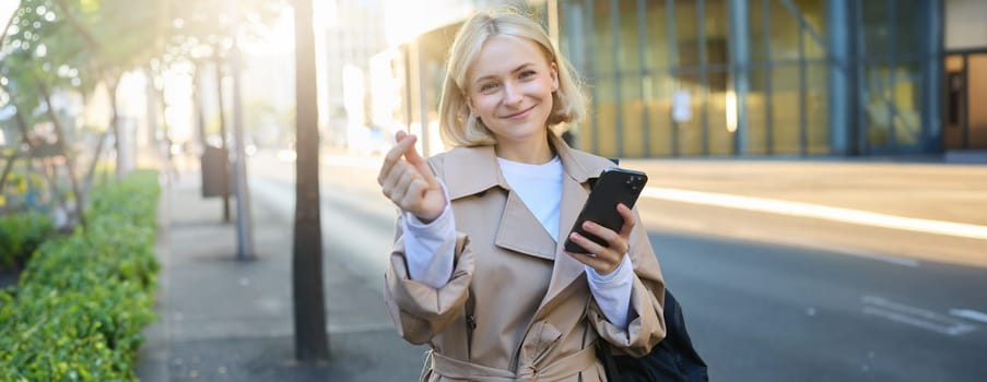 Lovely young female model in trench coat, showing heart gesture with fingers, holding smartphone and smiling at camera, standing on empty sunny street.