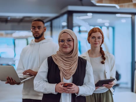 A diverse group of young businessmen, including a Muslim woman wearing a hijab, an orange-haired woman, and an African American, stand together in a modern office, showcasing a vibrant and inclusive workspace.