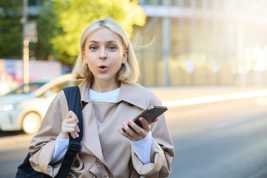 Image of young surprised woman, standing on street with mobile phone, looking amazed at camera, saying wow.