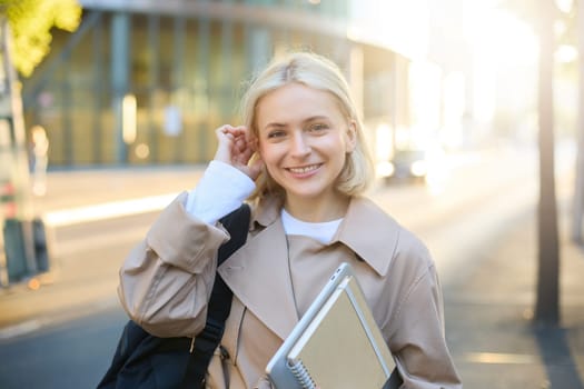 Young blonde woman, college student on street, holding journals and notebook, carry her homework material, going to university, smiling and looking happy.
