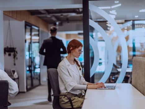 A young businesswoman with orange hair sitting confidently, fully engaged in her work on the laptop, exuding creativity, ambition, and a vibrant sense of individuality.