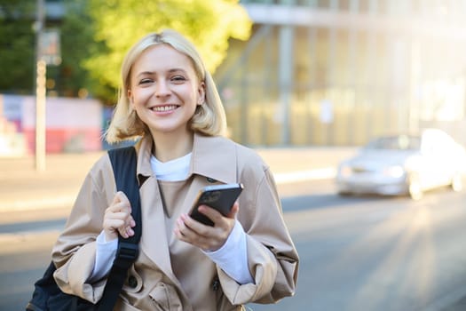 Close up portrait of happy, beautiful young woman with backpack, standing on street and using mobile phone, waiting for taxi near road, smiling.