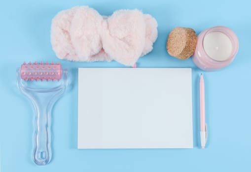 Blank notebook, wooden numbers 2022, pink massager, candle, fluffy headband and pen lie on a blue background with copy for text, close-up top view. Body care, beauty concept.