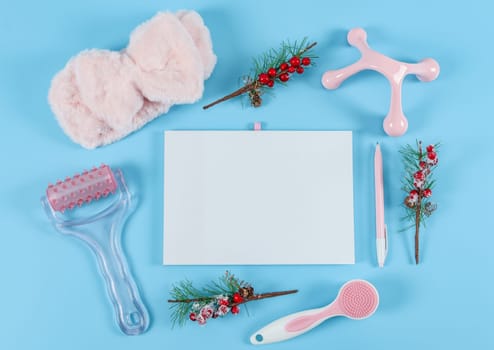 Blank notebook, wooden numbers 2022, fir branches, pink massager, candle, fluffy headband and pen lie on a blue background with copy for text, close-up top view. Body care, beauty concept.