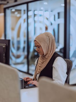 In a modern office, a young Muslim entrepreneur wearing a hijab sits confidently and diligently works on her computer, embodying determination, creativity, and empowerment in the business world